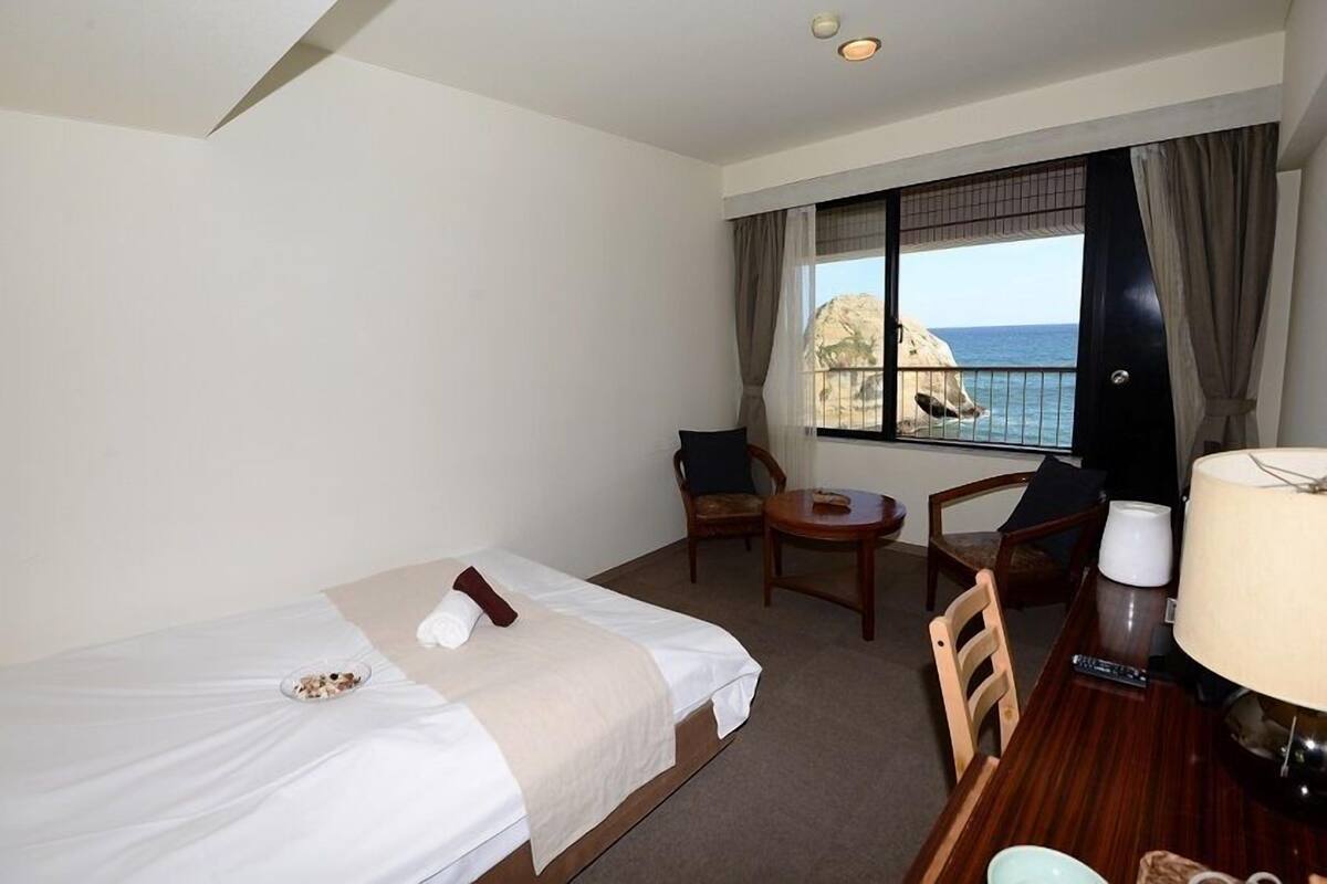 Een hotelkamer met een bed, een kleine tafel met een lamp, een stoel en uitzicht op zee door een raam.