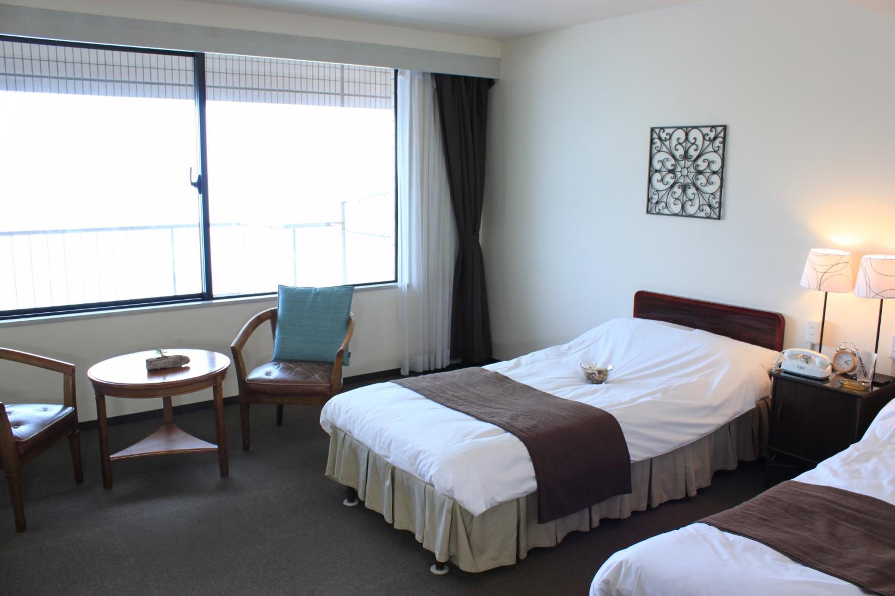 Habitación de hotel con dos camas, una mesita, una silla, una ventana con cortinas, una mesita de noche con lámpara y un adorno en la pared.