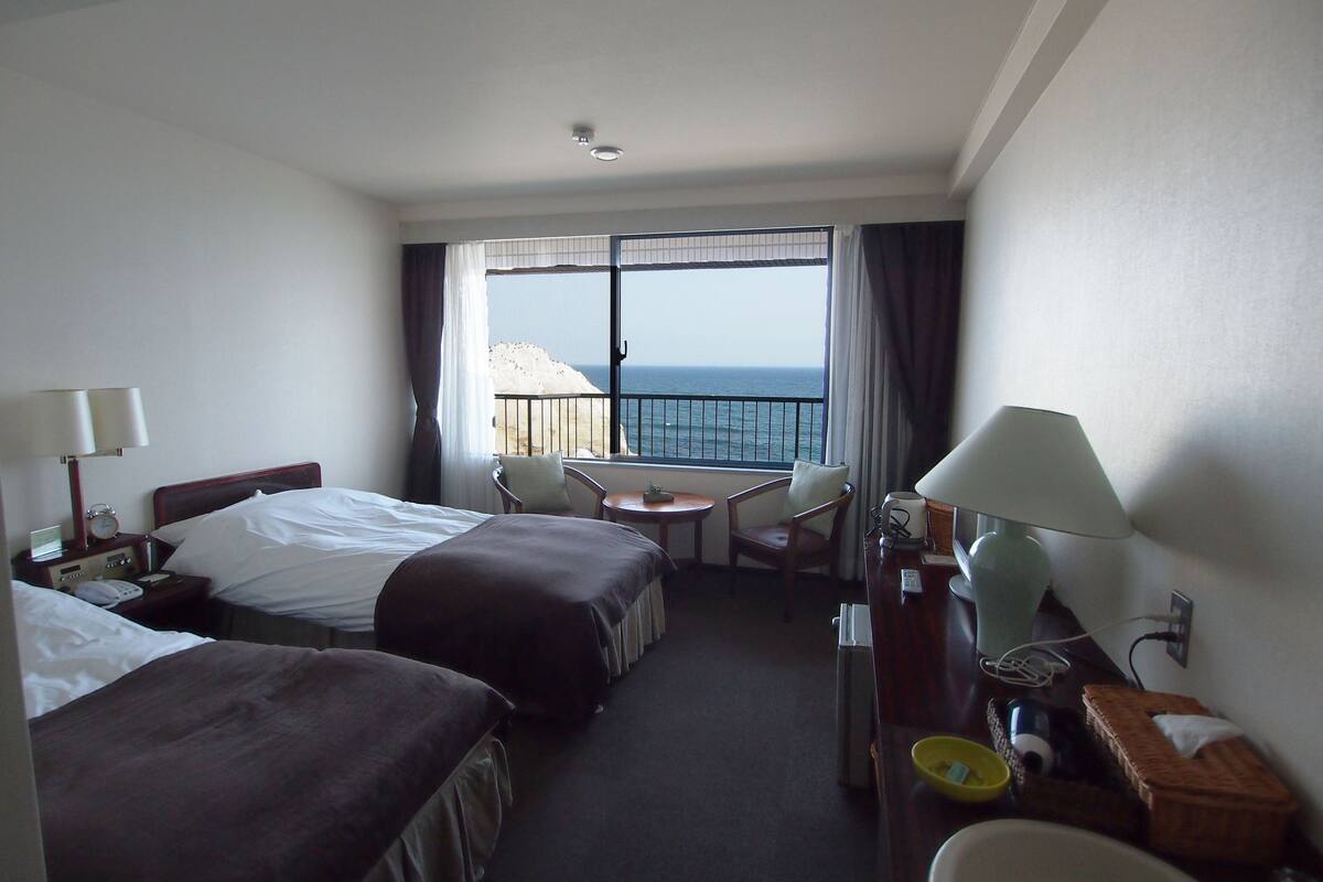 Een hotelkamer met twee bedden, een tafeltje, een stoel, een lamp en uitzicht op zee.