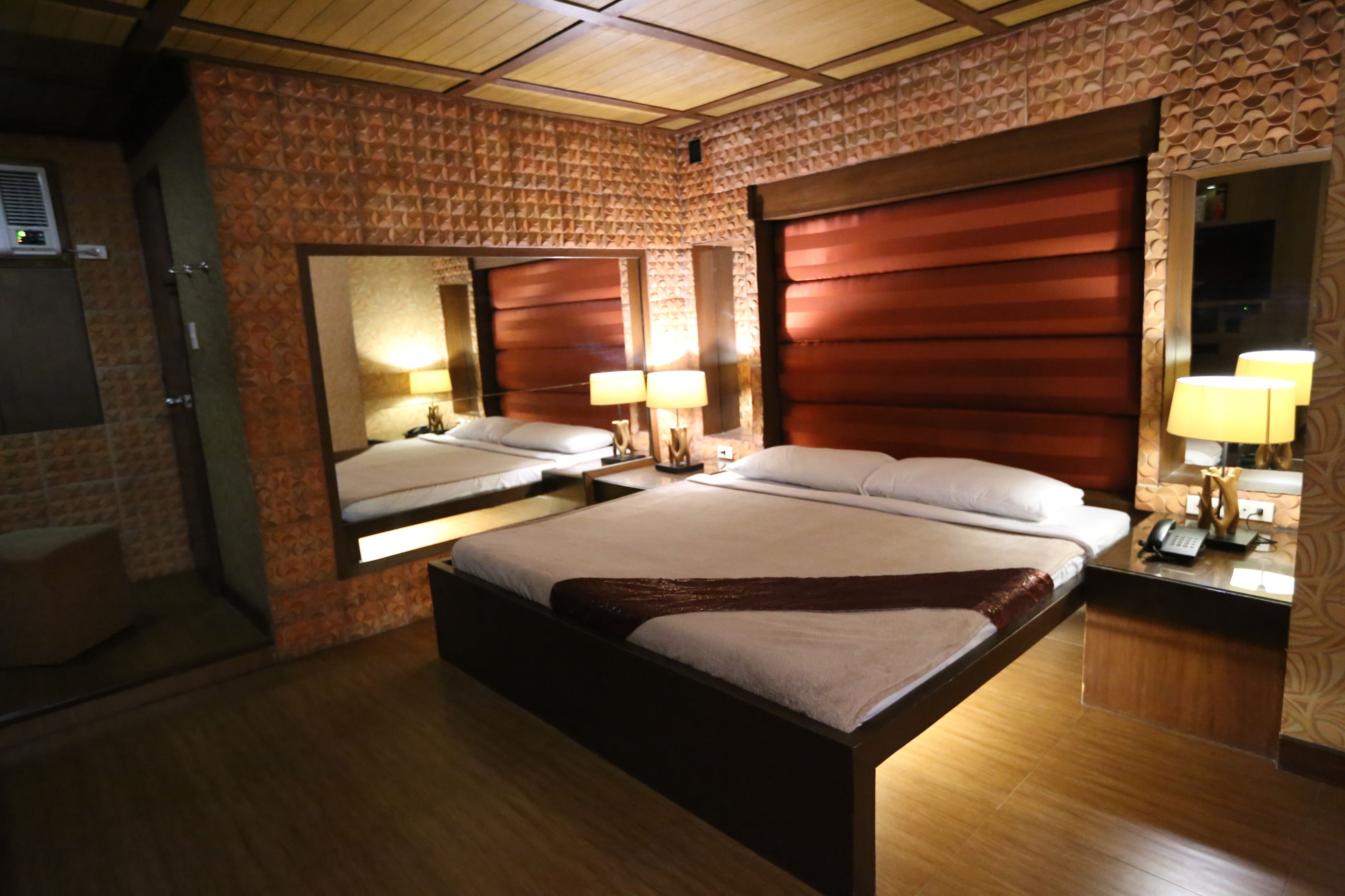 A hotel room with two beds, a wooden floor, a large mirror, and a bedside table with a lamp.