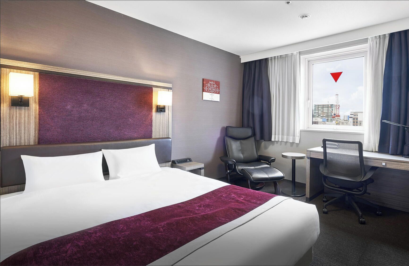 Une chambre d’hôtel avec un grand lit, un bureau, une chaise, une petite table, une lampe et une fenêtre donnant sur la ville.
