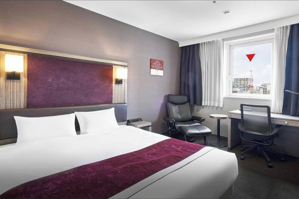 Une chambre d’hôtel avec un grand lit, un bureau, une chaise, une petite table, une lampe et une fenêtre donnant sur la ville.