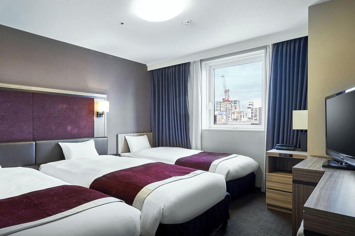 A hotel room with two beds, a desk, a TV, and a window with a city view.