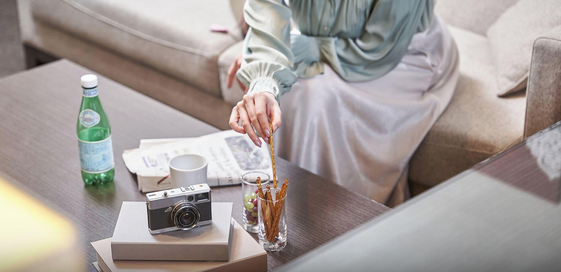 A person sitting on a couch, holding a camera, with a bottle, glasses, and a box on the table.