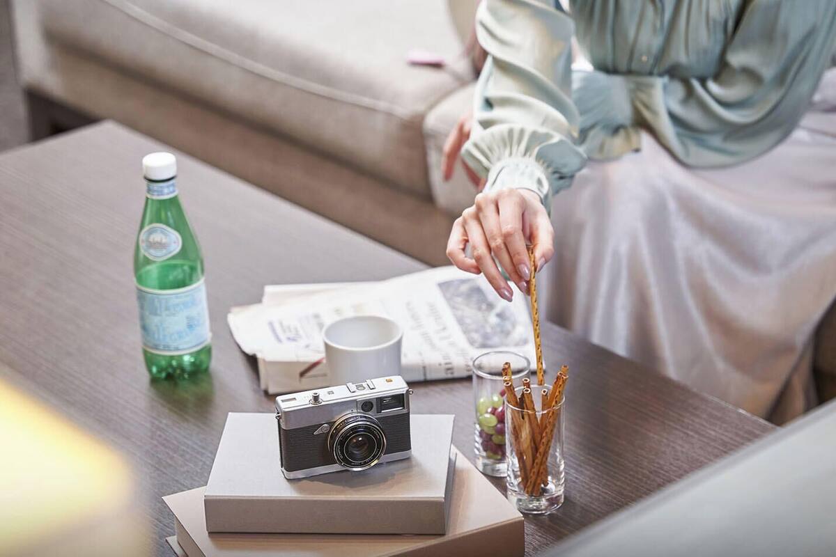 Eine Person sitzt auf einem Sofa, hält eine Kamera, und auf dem Tisch befinden sich eine Flasche, Gläser und eine Schachtel.