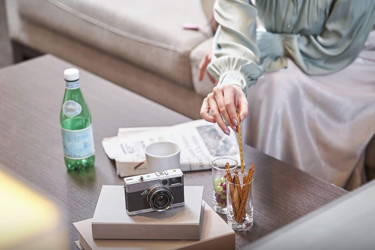 A person sitting on a couch, holding a camera, with a bottle, glasses, and a box on the table.
