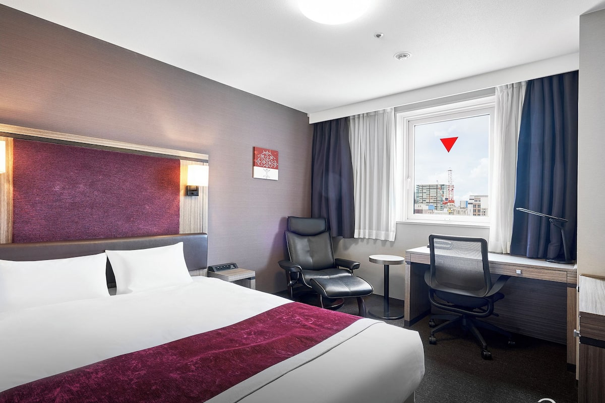 Ein Hotelzimmer mit einem großen Bett, einem Schreibtisch, einem Sessel und Blick auf die Stadt.