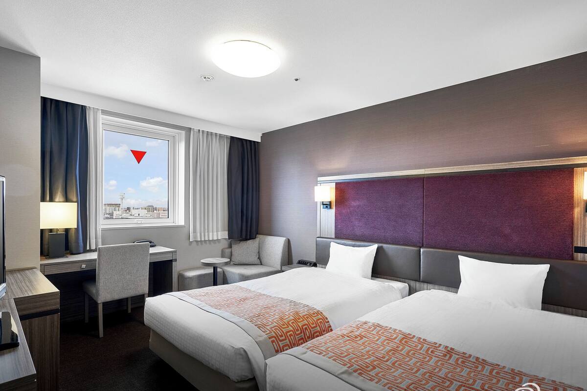 Ein Hotelzimmer mit zwei Betten, einem Schreibtisch, einem Sessel, einem Fenster mit Vorhängen und Blick auf die Stadt.