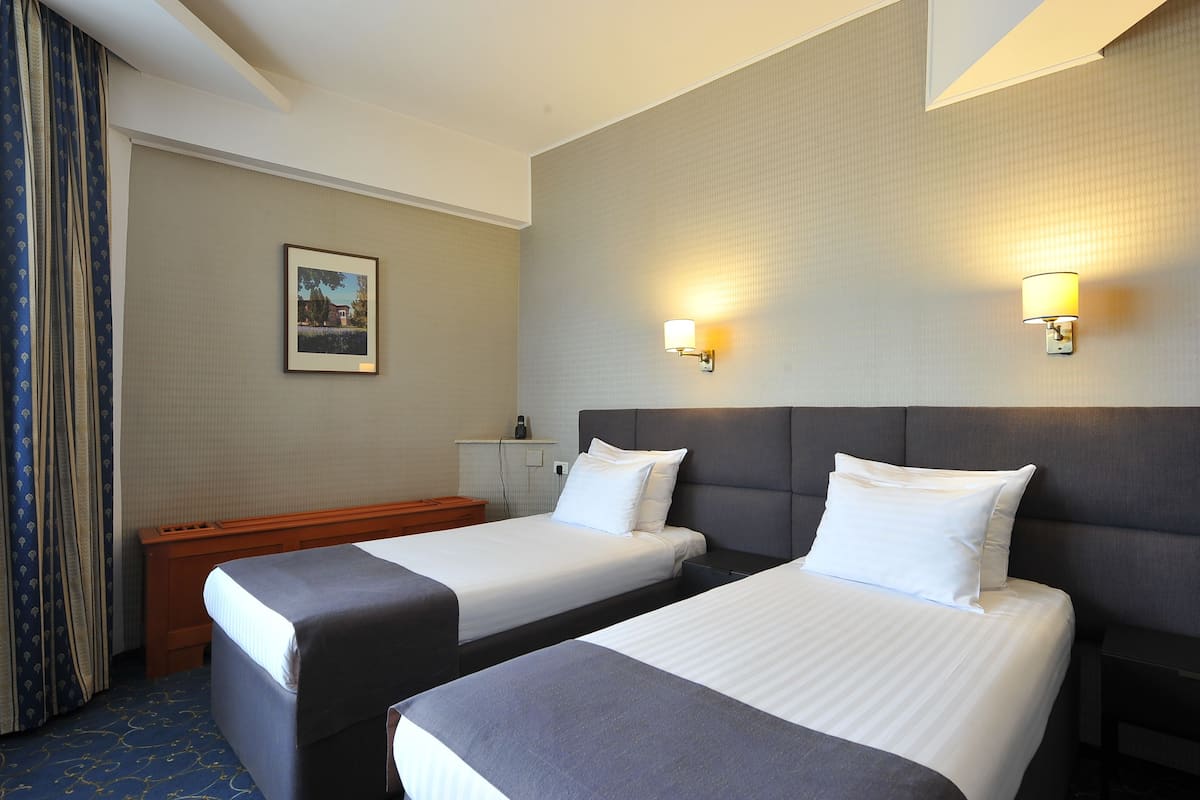 A hotel room with two beds, a headboard, a nightstand, a lamp, a framed picture on the wall, and a window with curtains.