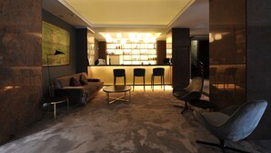 Un área de lounge moderna con un sofá, sillones y una mesita. Se observa una barra con estantes llenos de botellas al fondo.