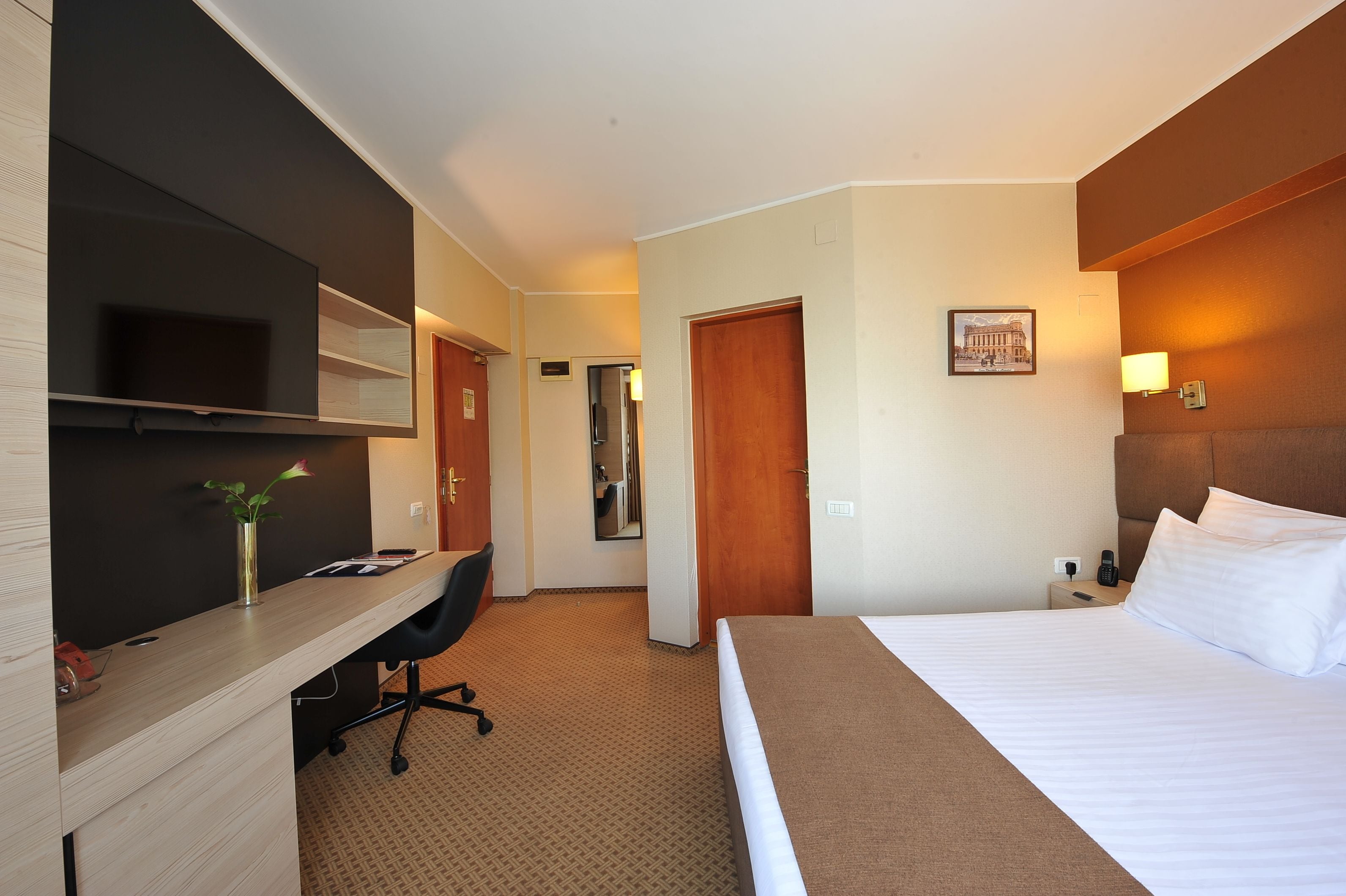 A hotel room with a bed, a desk with a chair, a flat-screen TV, a vase with flowers, a framed picture on the wall, and a nightstand with a lamp.