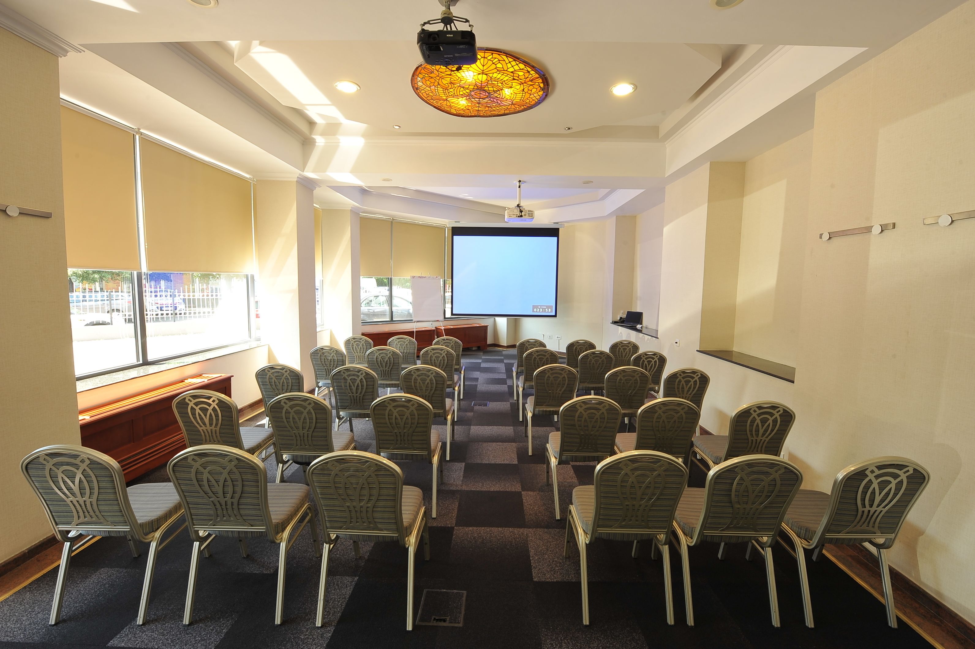 Un salón de conferencias con hileras de sillas orientadas hacia una pantalla grande, un proyector y una ventana con persianas.