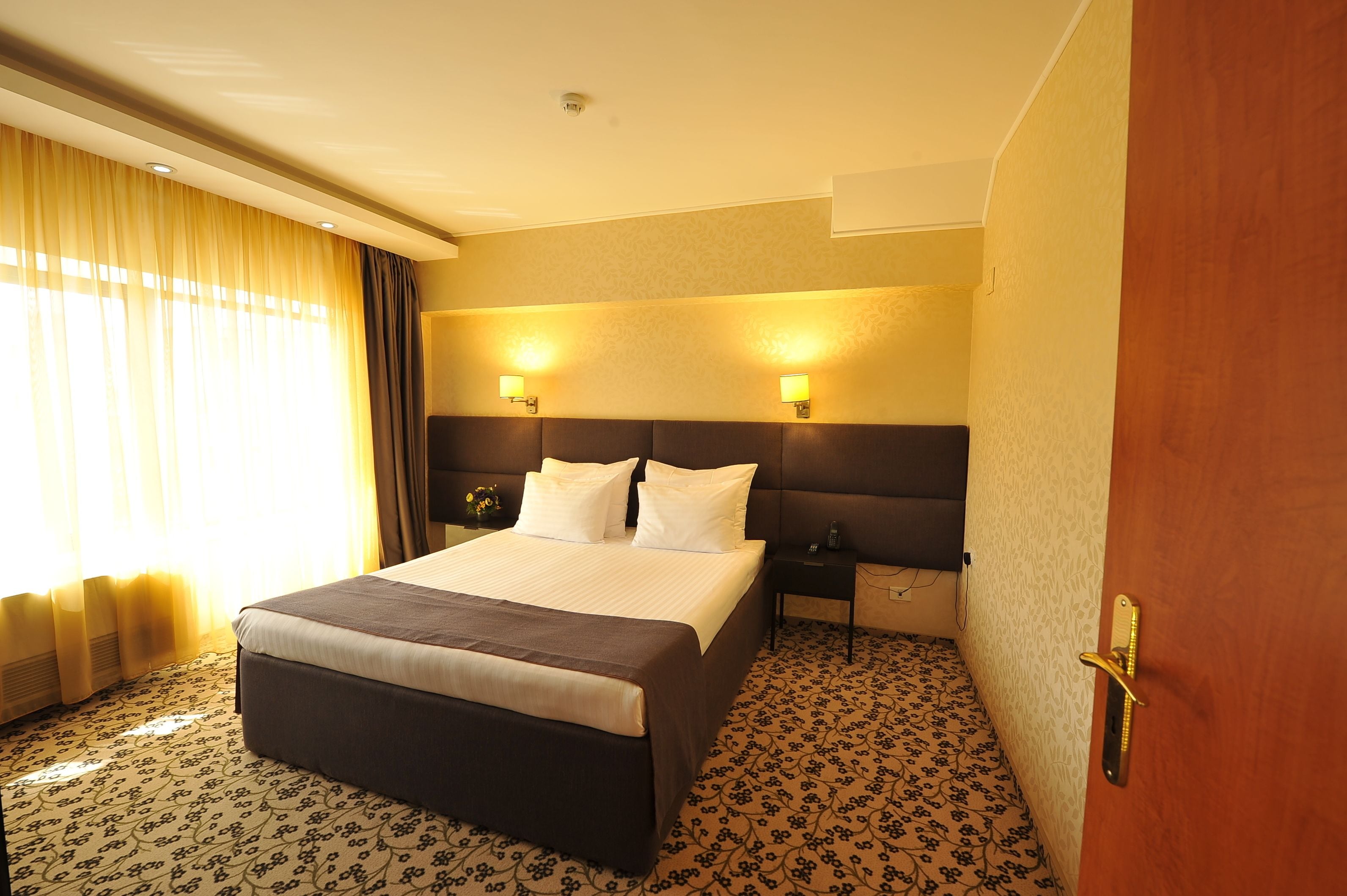A hotel room with a large bed, two bedside tables with lamps, a wooden door, and patterned carpet.