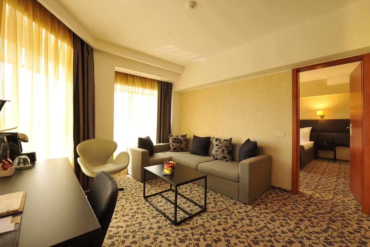A hotel room with a sofa, armchair, coffee table, and a desk with a lamp.