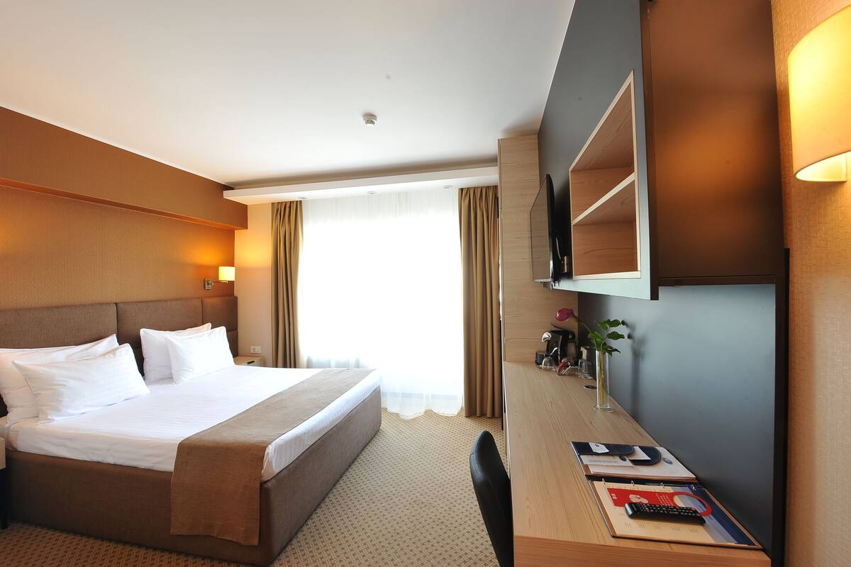 A hotel room with a large bed, a desk with a chair, a TV, and a window with curtains.
