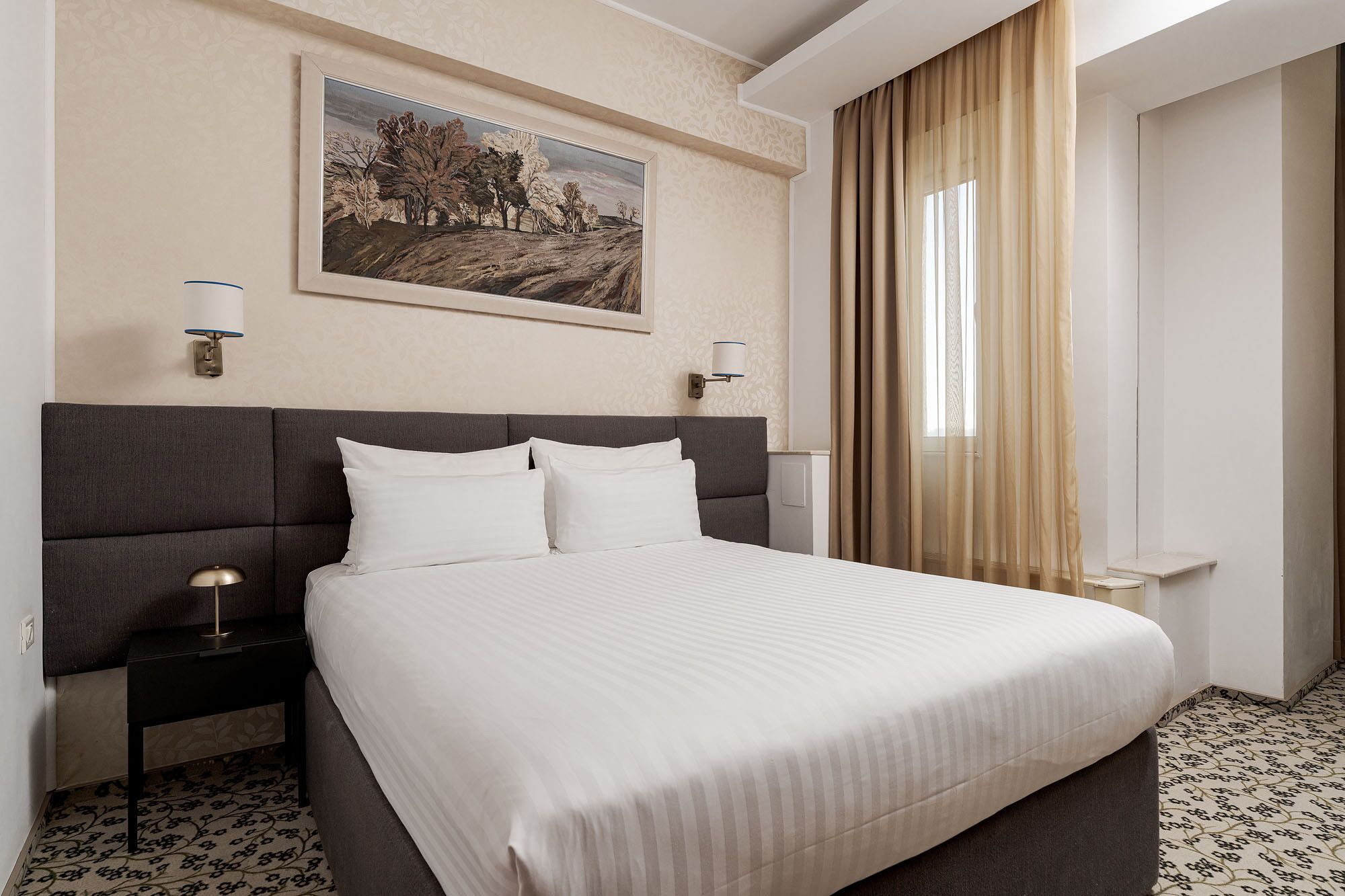Quarto de hotel com uma cama grande, duas mesas de cabeceira, um criado-mudo, um quadro na parede e uma janela com cortinas.