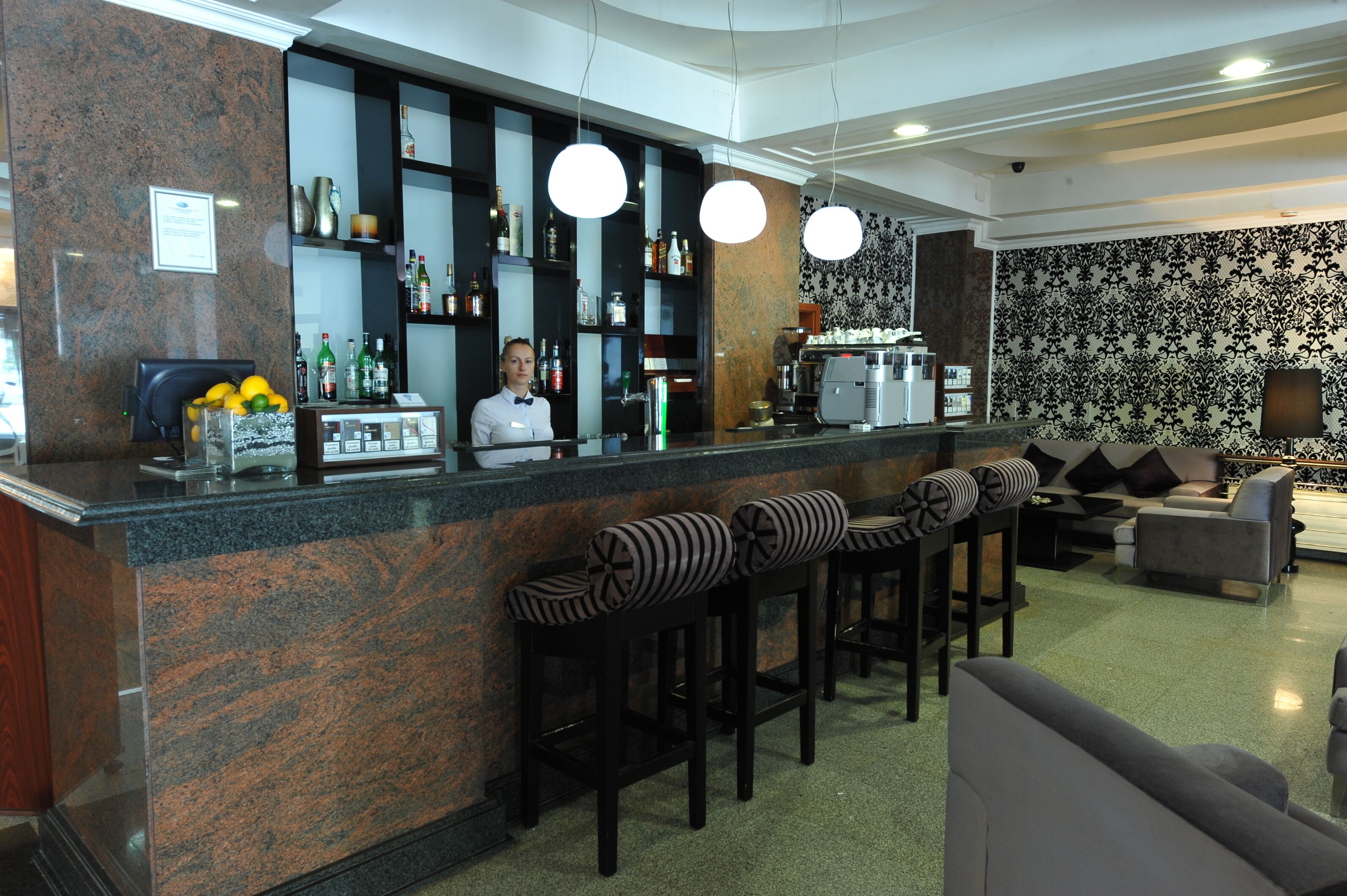 Una zona de bar con mostrador, taburetes y un barista. Hay estanterías con botellas y una máquina de café. La habitación tiene un papel tapiz estampado y un sofá.
