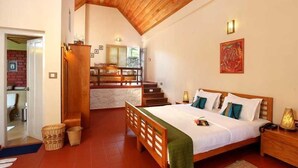 A bedroom with a wooden bed, a nightstand, a lamp, and a view of a kitchen and living area through an open door. - Ambady Estate (Devikolam)