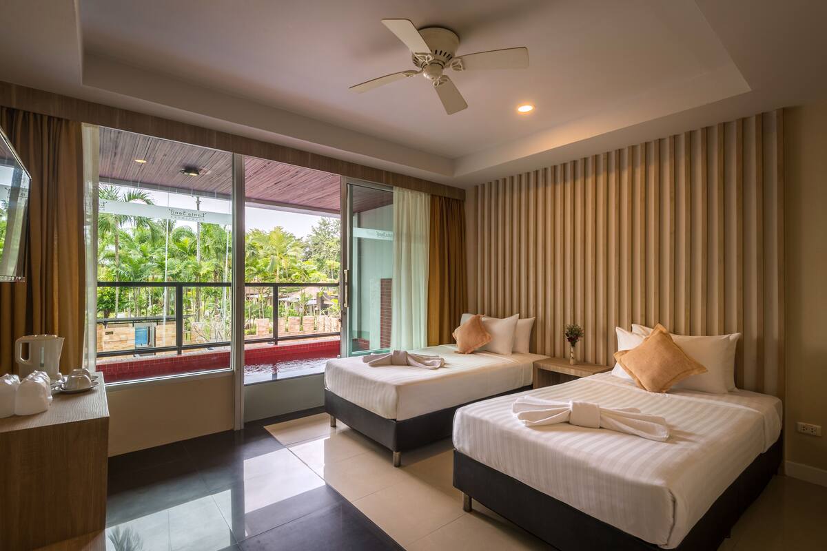 A hotel room with two beds, a ceiling fan, a large window with a view of greenery, and a sliding glass door.