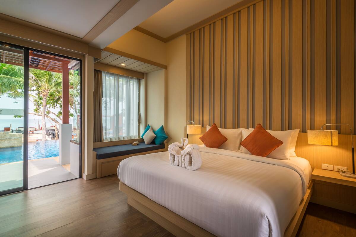 A hotel room with a large bed, a bedside table with a lamp, a window with curtains, and a view of a pool and palm trees.