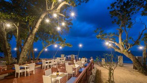 Un área de comedor al aire libre con mesas y sillas, rodeada de árboles y con vista a una playa.