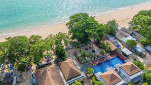 Vista aérea de un resort frente a la playa, con piscina, mobiliario en la arena y abundante vegetación.
