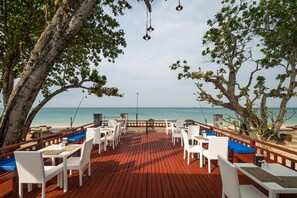 A wooden deck with white wicker furniture and blue cushions, overlooking a beach.