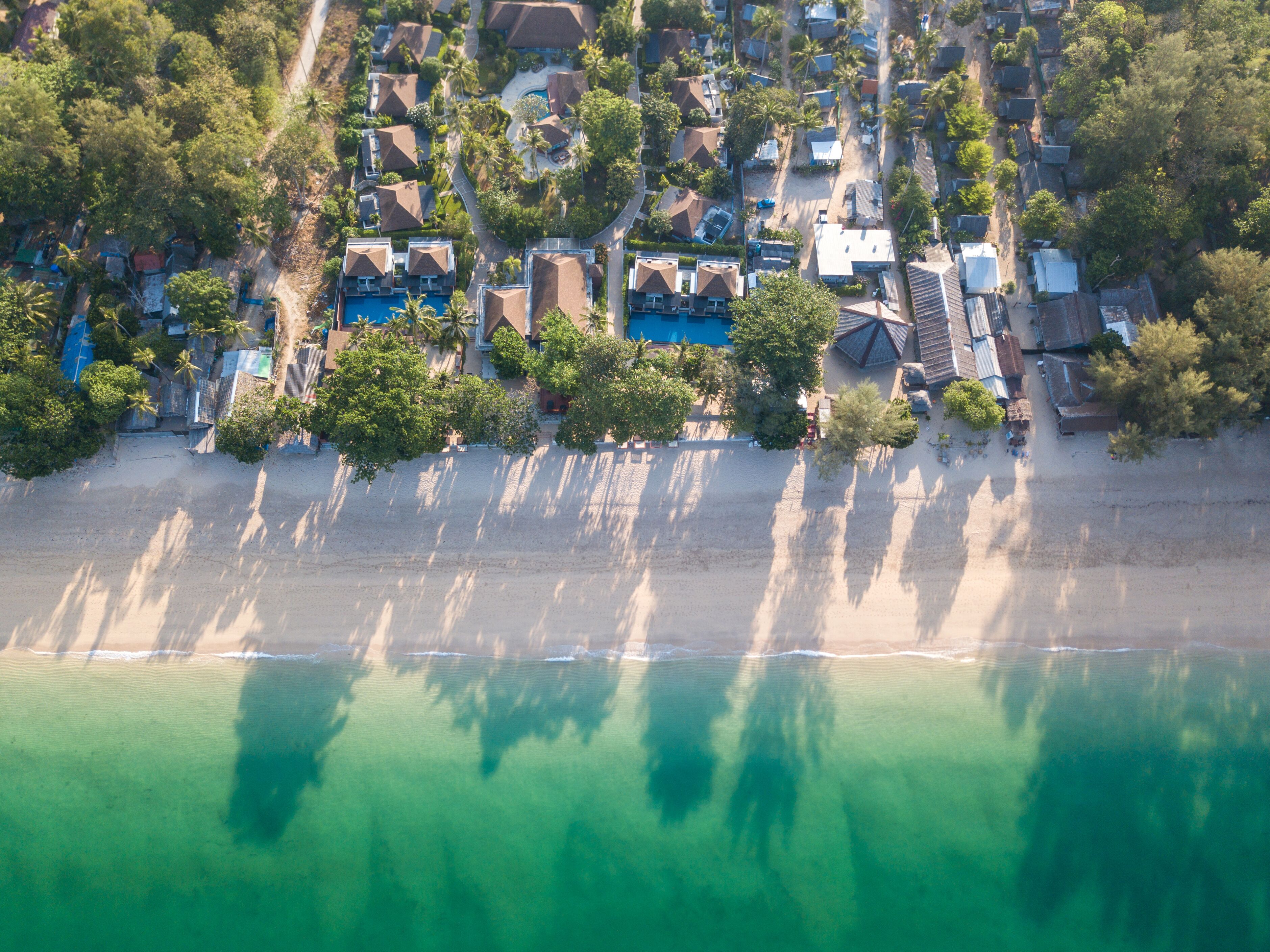 Vista aèria d'una zona residencial a la costa, amb cases, una platja i un mar blau clar.