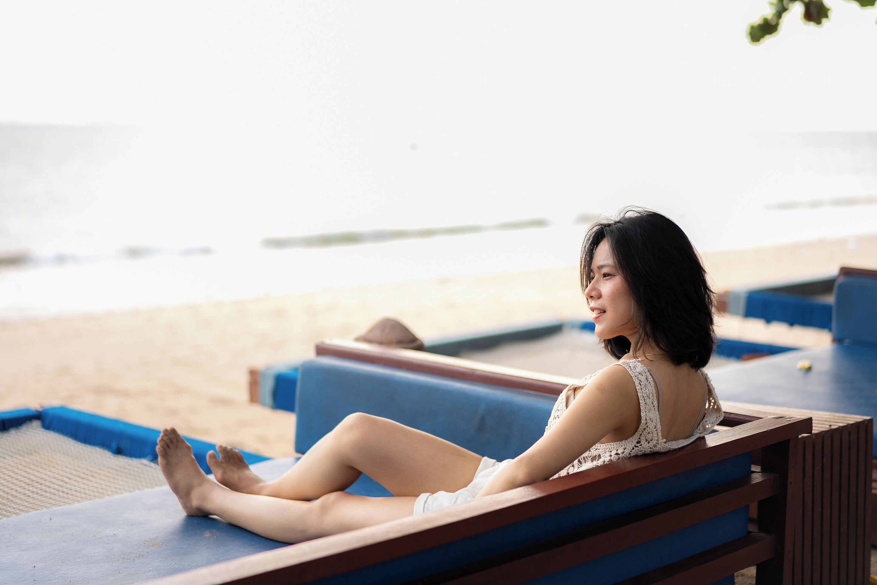 Une personne est assise sur un fauteuil de plage bleu, dont l’armature est en bois, et elle a devant elle une plage de sable bordée par l’eau.