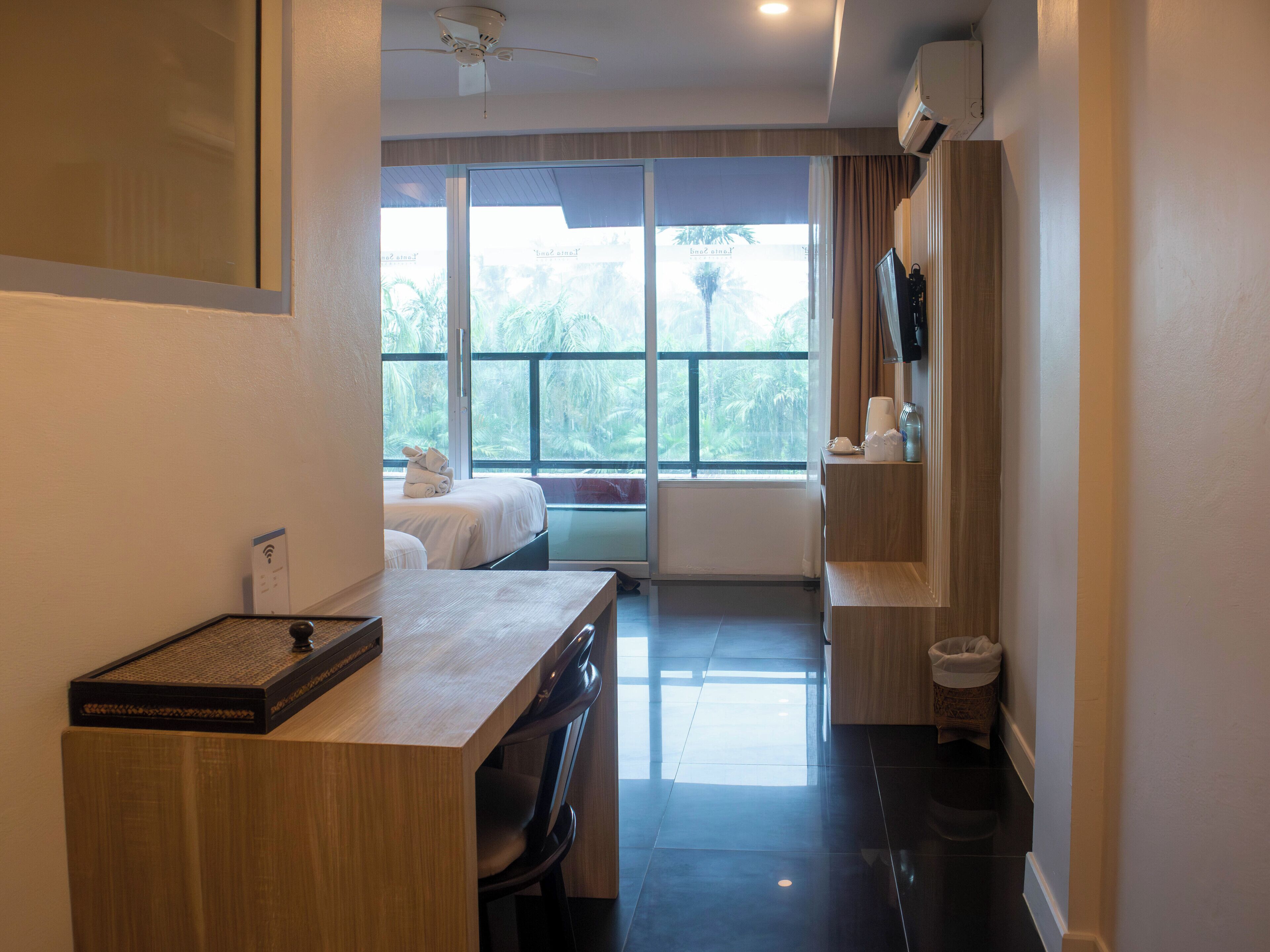 A hotel room with a desk, a bed, a TV, and a window with a view of palm trees.