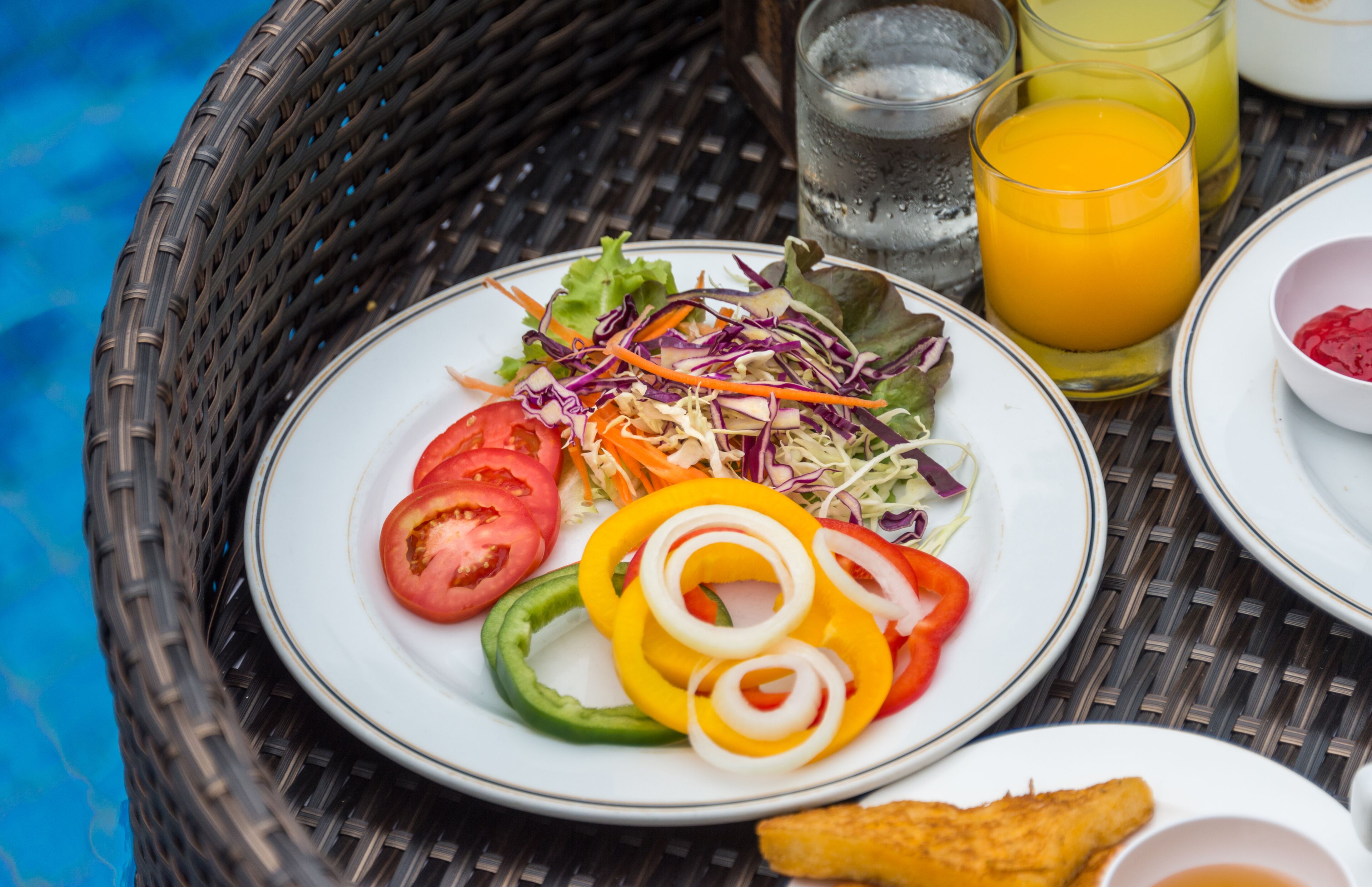 A wicker basket containing a plate of salad with sliced vegetables and a small bowl of sauce, a glass of orange juice, and a glass of water.