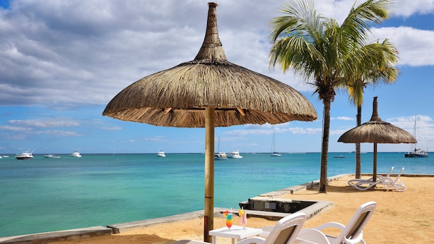 A beach with white lounge chairs, a thatched umbrella, and a view of the ocean with boats. - Coral Azur Beach Resort (Mont Choisy)
