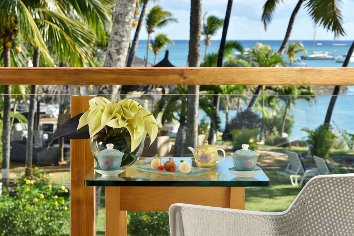 A glass table with a vase of flowers, a teapot, and a teacup on a balcony overlooking a beach with palm trees and boats.