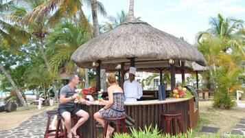 A thatched-roof outdoor bar with a chef, a man and a woman seated.
