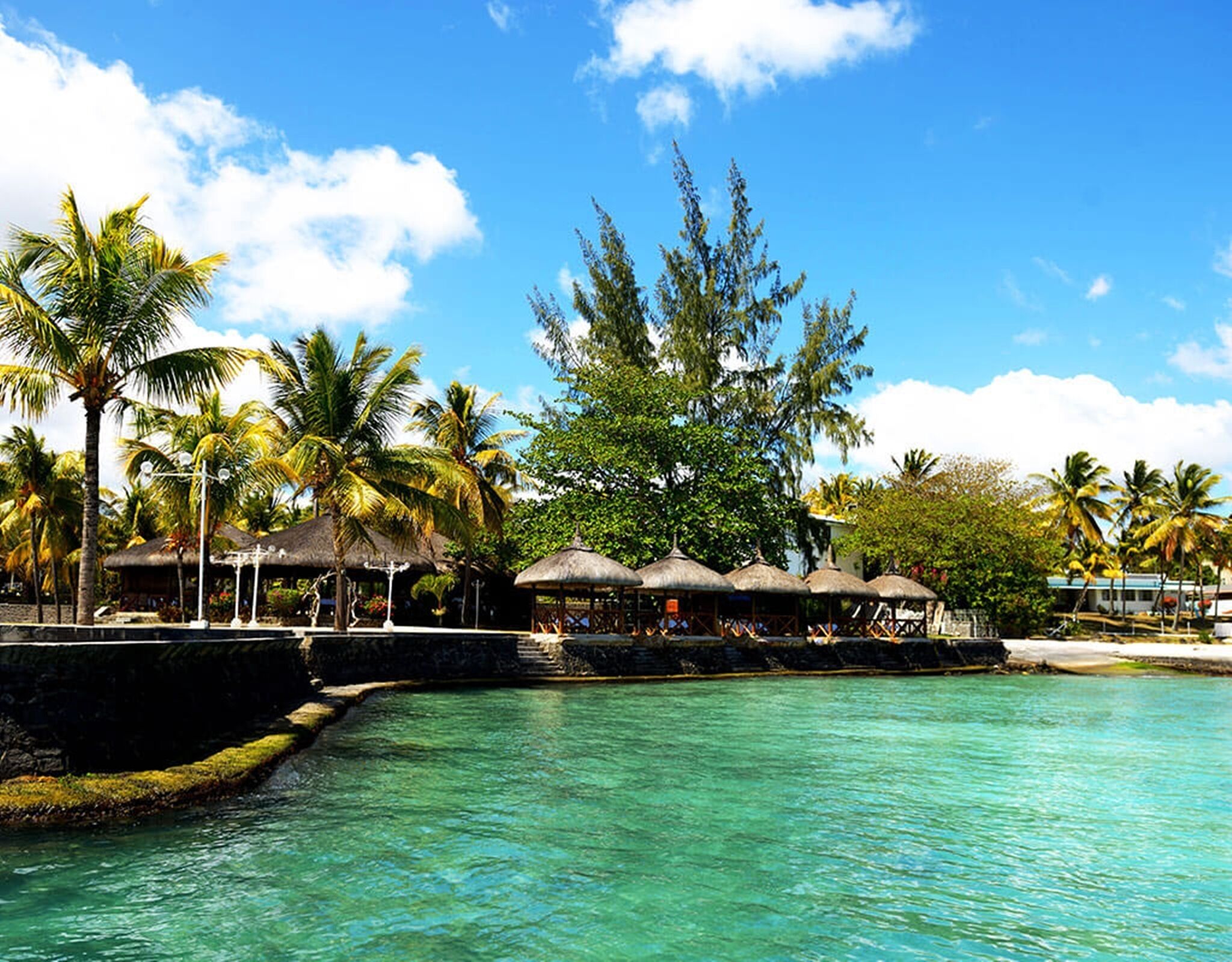 A tropical resort with clear turquoise water, palm trees, and thatched-roof structures.