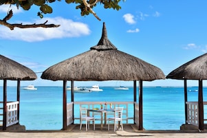 A beachside view with thatched umbrellas and a table with chairs, overlooking the sea and boats.