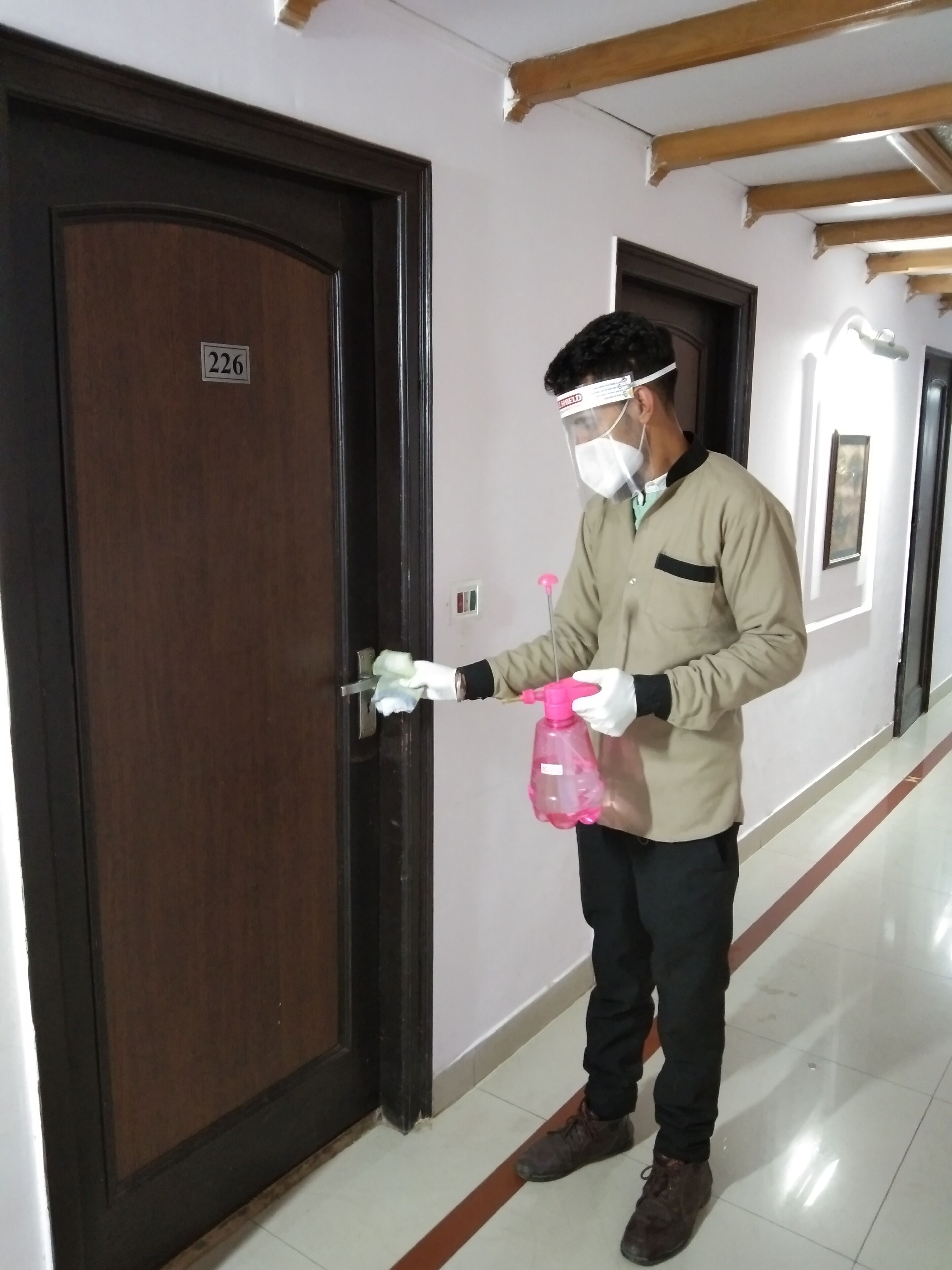 A person in a beige jacket and gloves holding a spray bottle, standing in a hallway with a wooden door marked "226".
