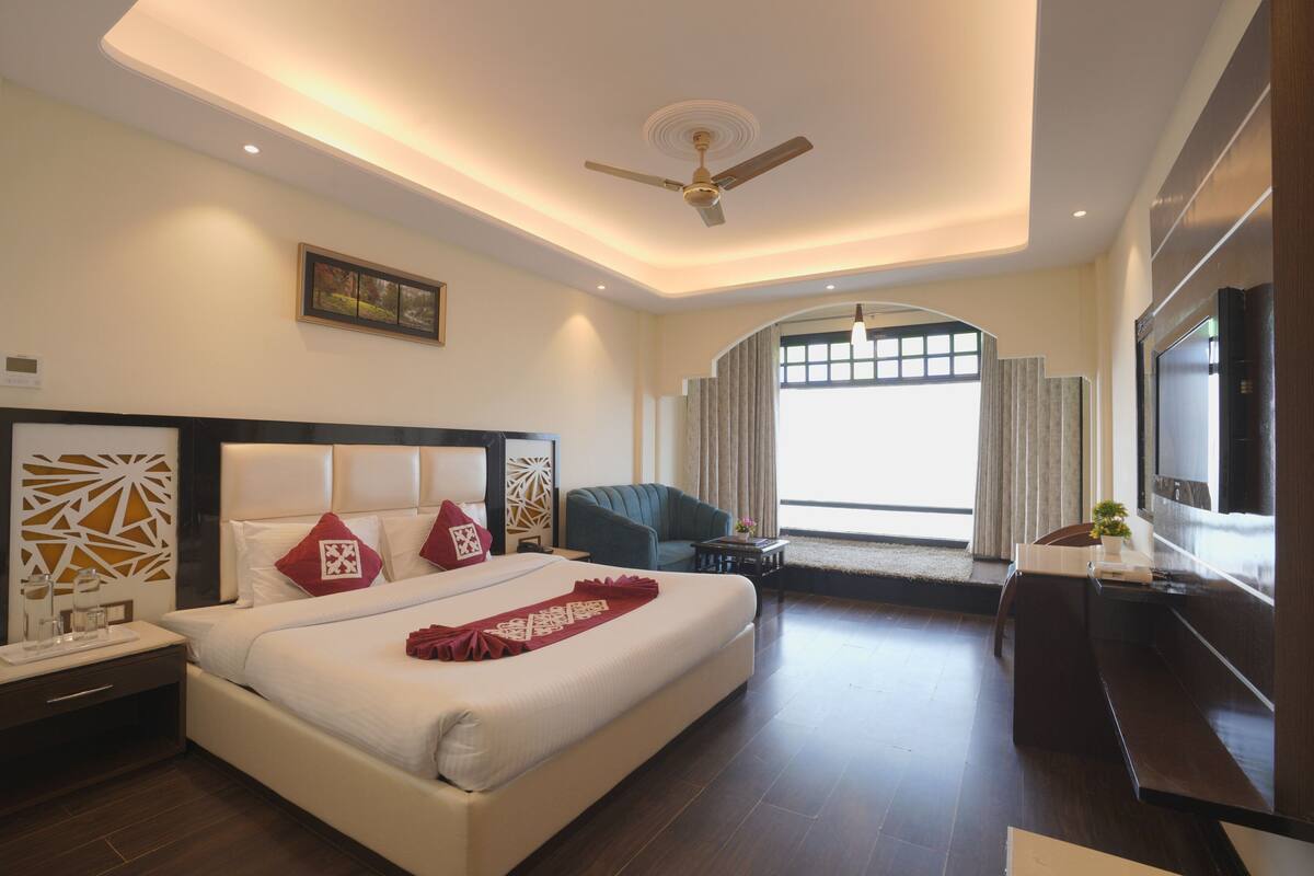 A hotel room with a large bed, a ceiling fan, a TV, a seating area with a chair, and a window with curtains.