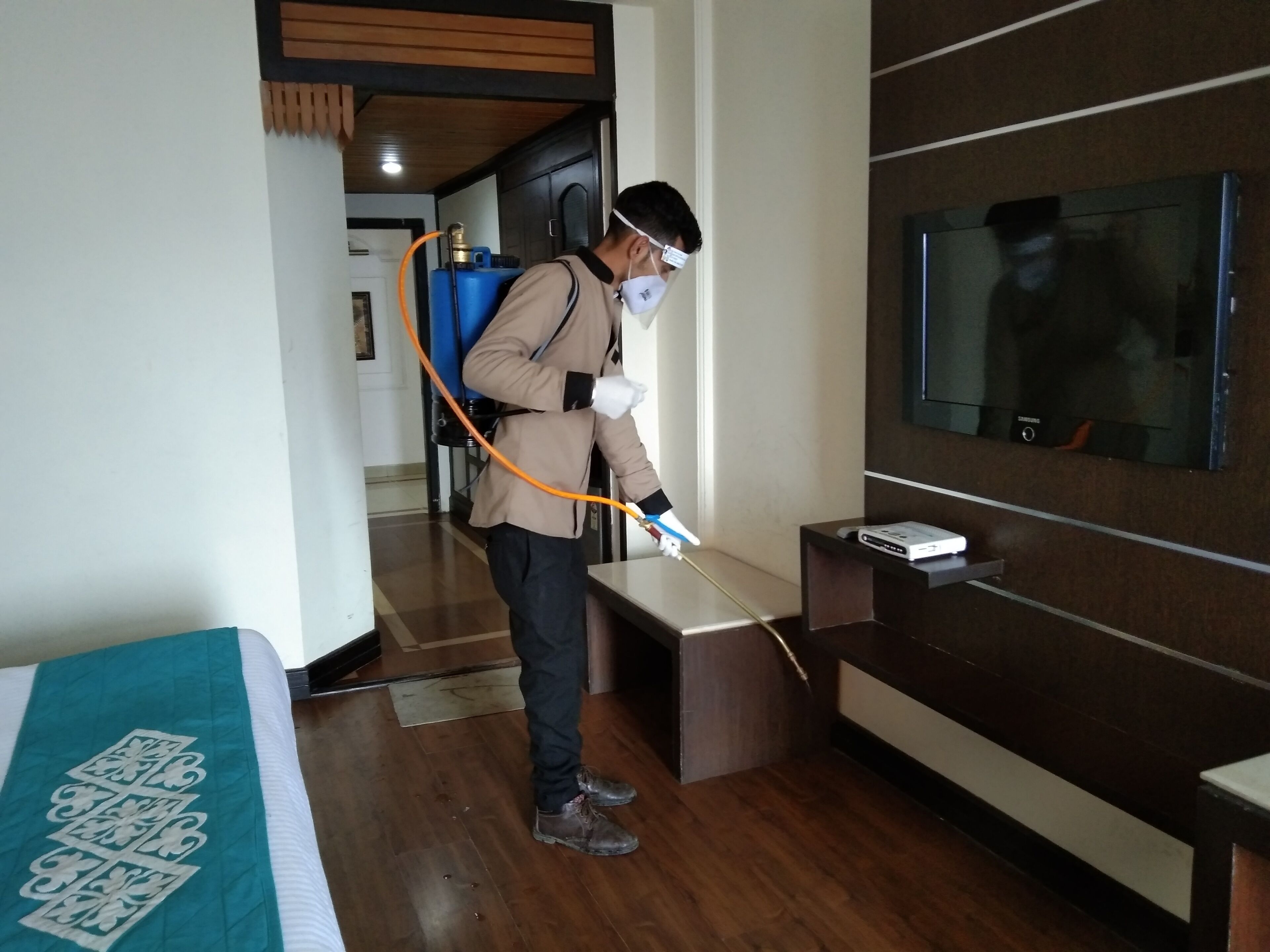 A person using a spray device in a hotel room with a wooden floor, a bed with a teal bedspread, and a flat-screen TV mounted on the wall.