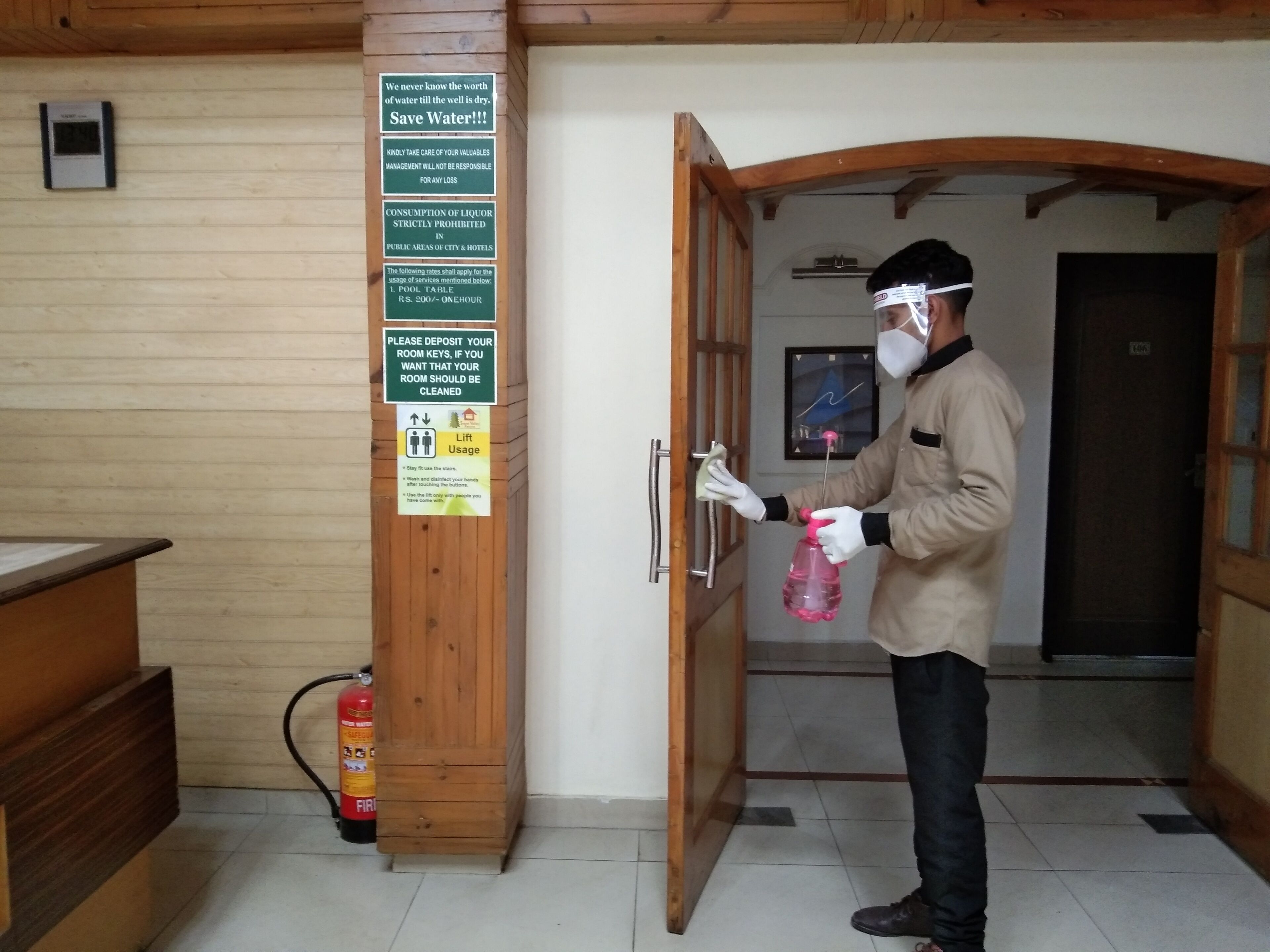 A person cleaning a door handle with a spray bottle, a fire extinguisher, and a sign about water conservation and room key deposit.