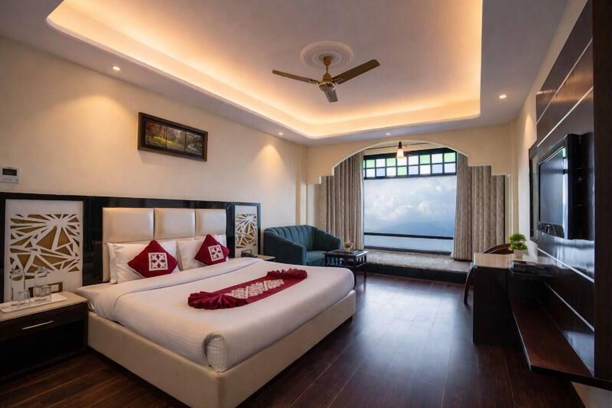 A modern hotel room with a large bed, a ceiling fan, a TV, and a view of the city through the window.
