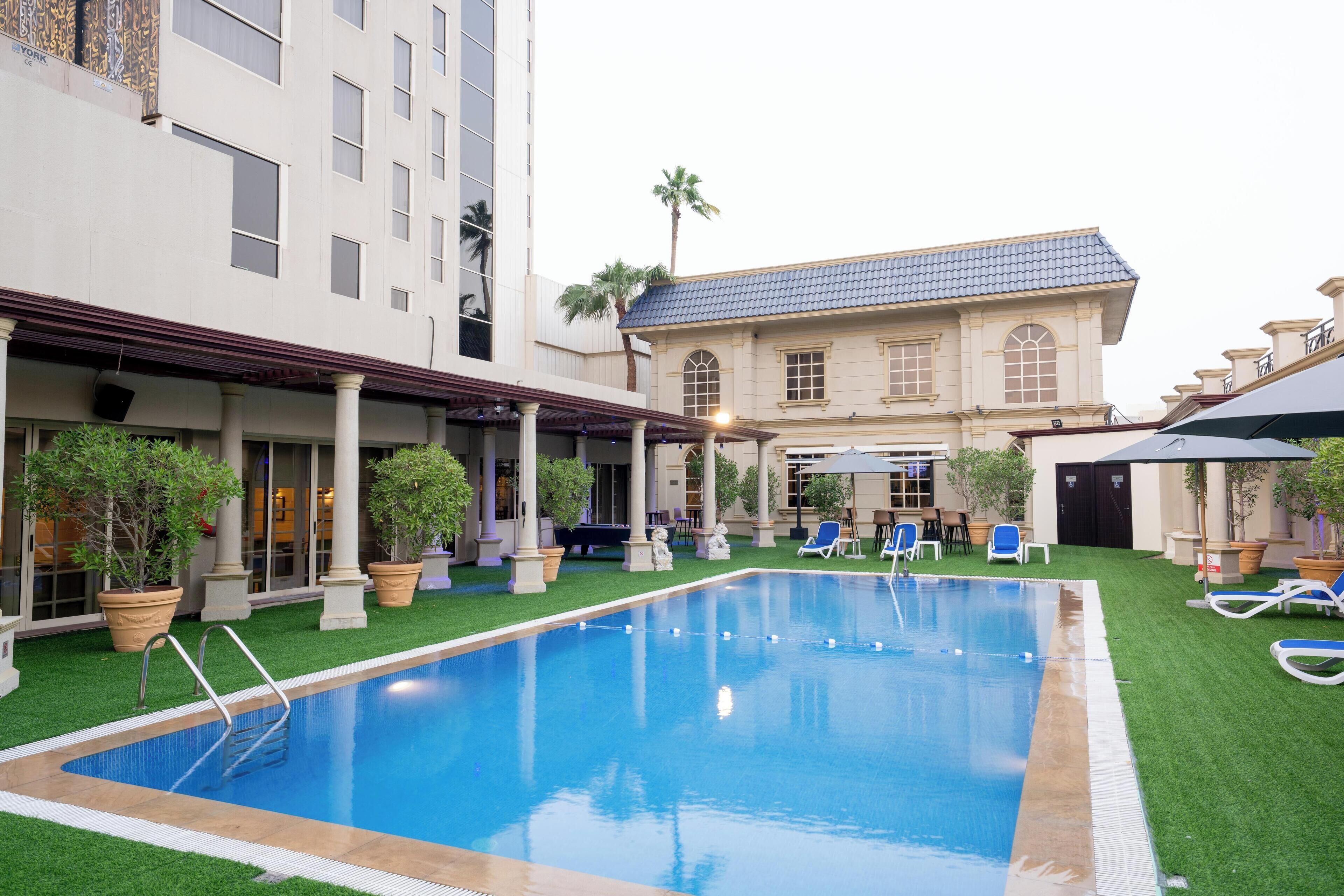 Un espace piscine d’hôtel comprenant une piscine rectangulaire, des chaises longues et un bâtiment doté de colonnes et d’un toit recouvert de tuiles.