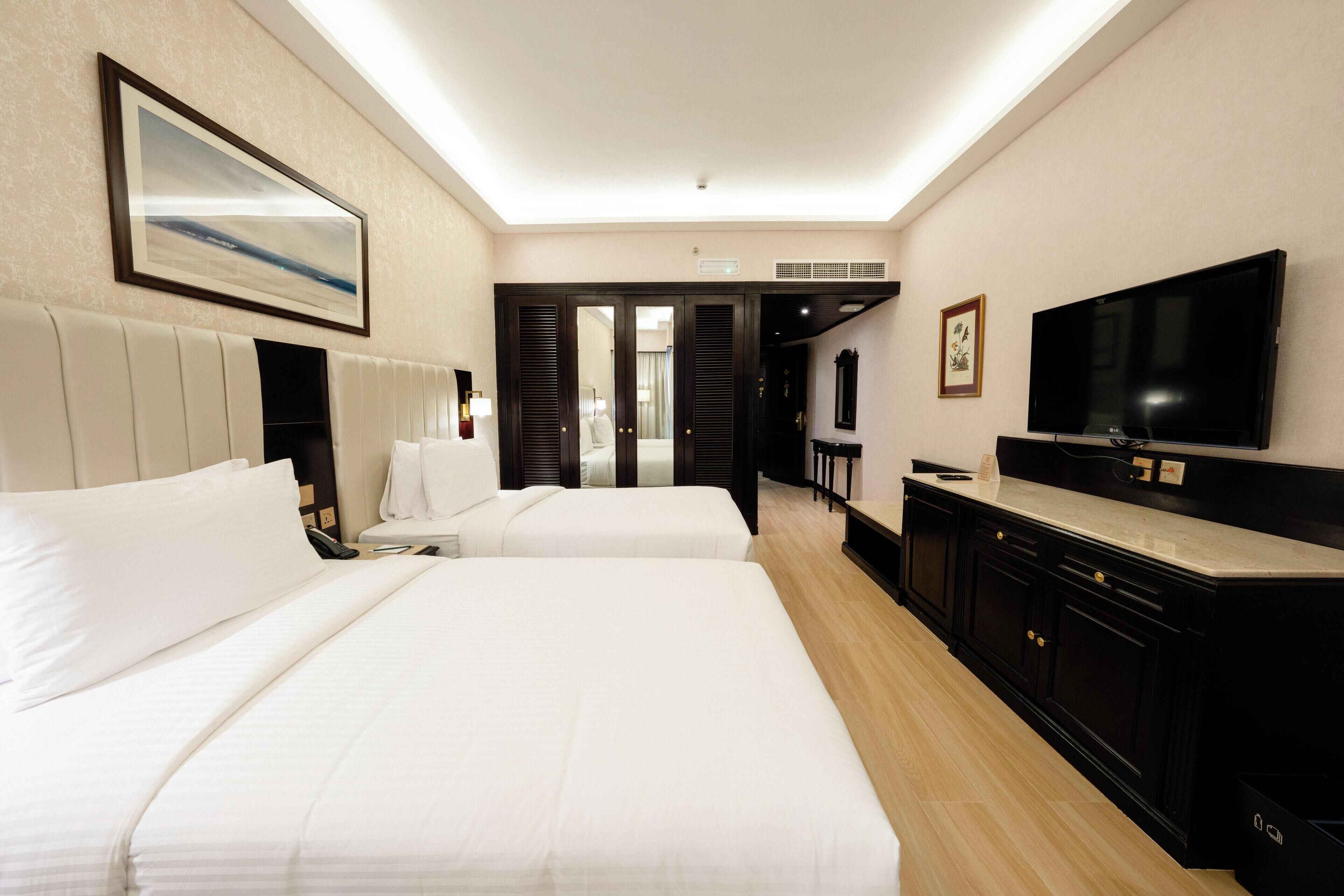 Habitación de hotel con dos camas, televisor, escritorio y baño visible a través de una puerta abierta.
