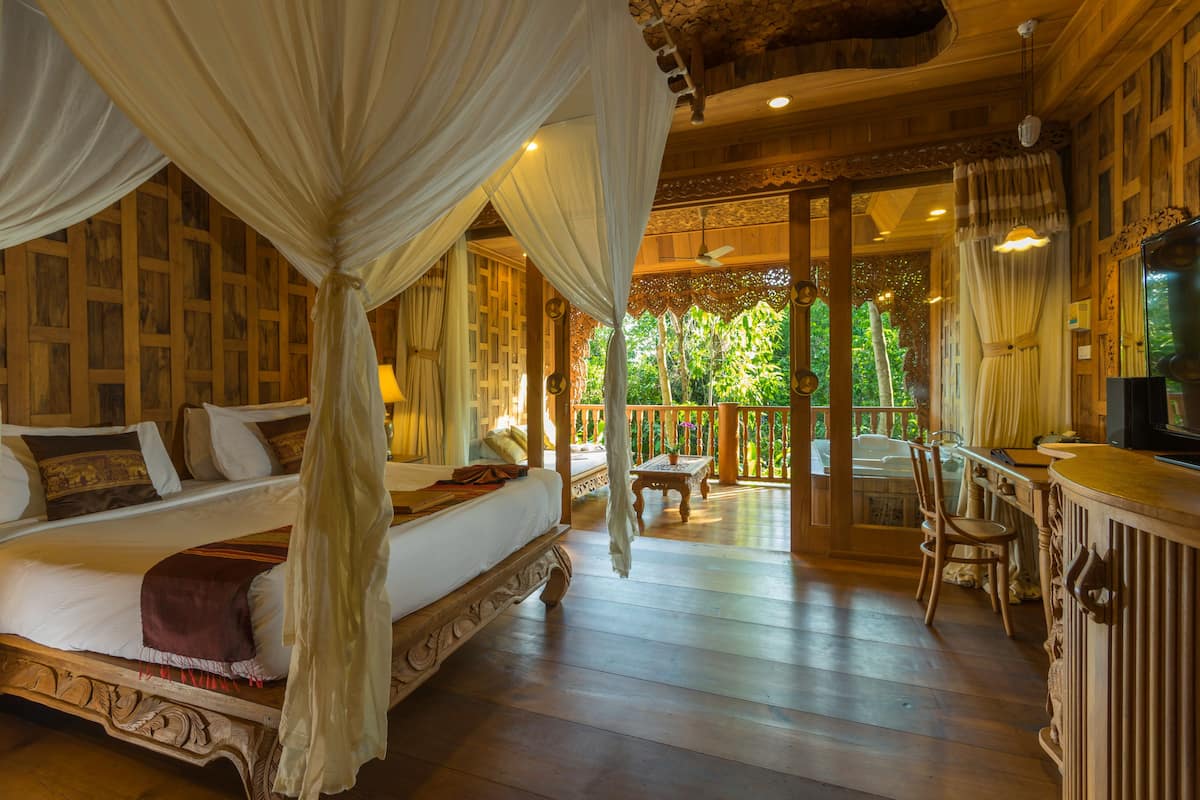 A bedroom with a four-poster bed, a wooden desk, and a balcony with a view of greenery.