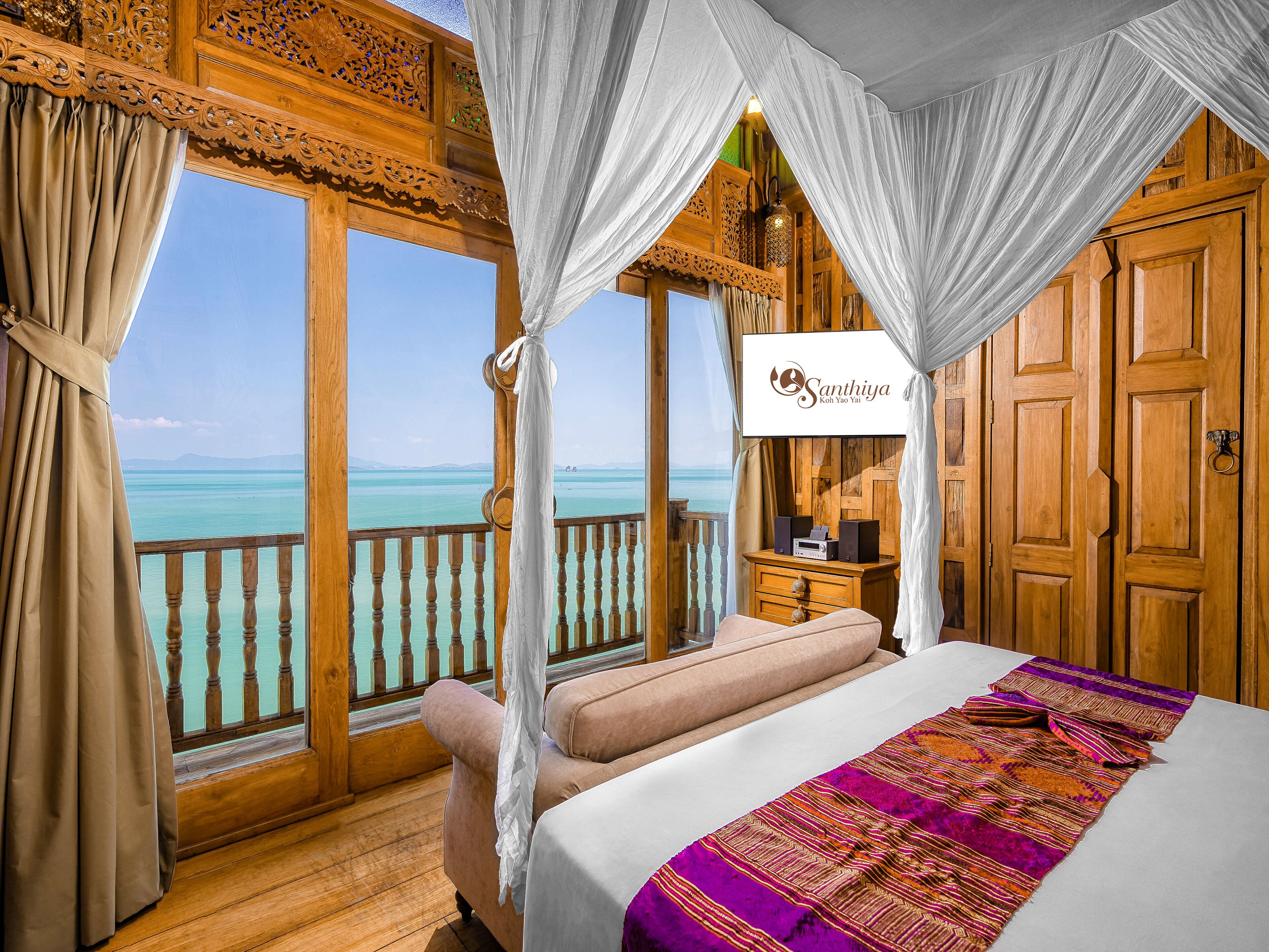 Ein Schlafzimmer mit großem Fenster und Blick auf ein Gewässer, einem Bett mit bunter Decke und Holzmöbeln.