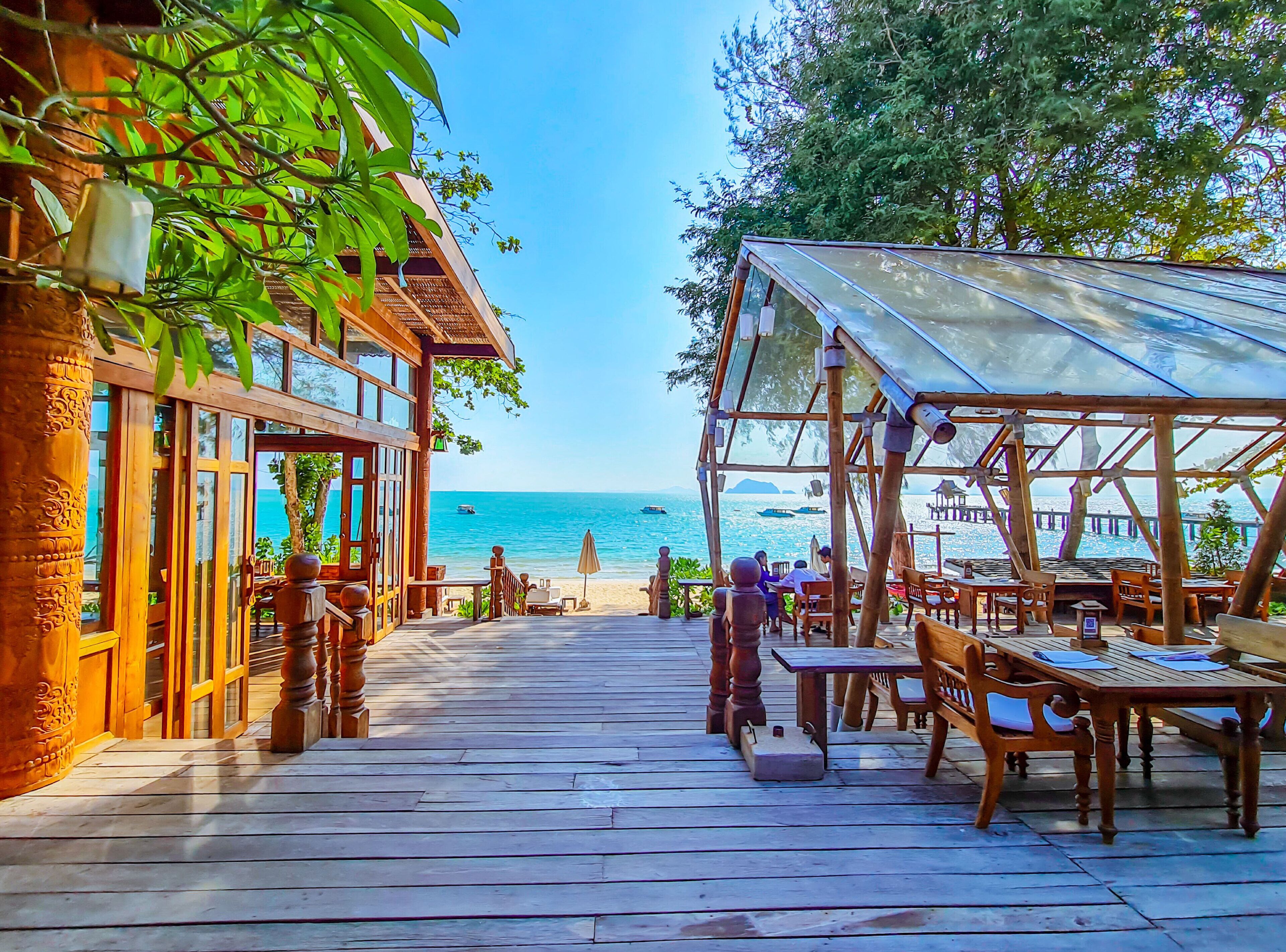 Una terraza de madera con mesas y sillas, con vista a la playa y al mar.