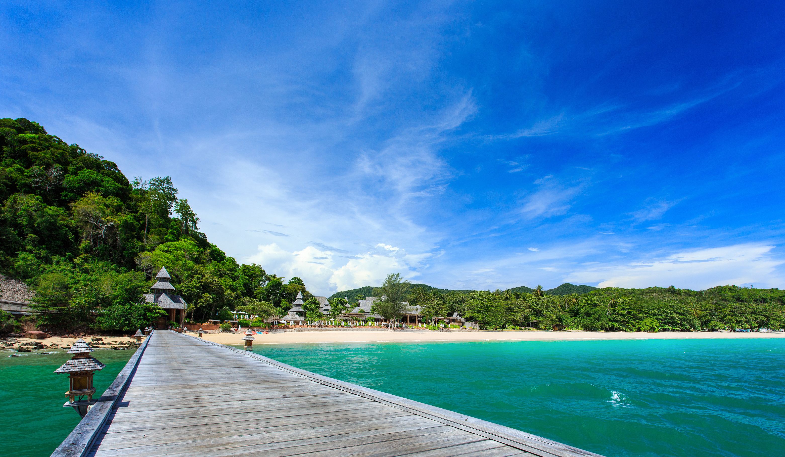 A wooden pier extending into turquoise waters, leading to a beach with lush greenery and traditional-style buildings.