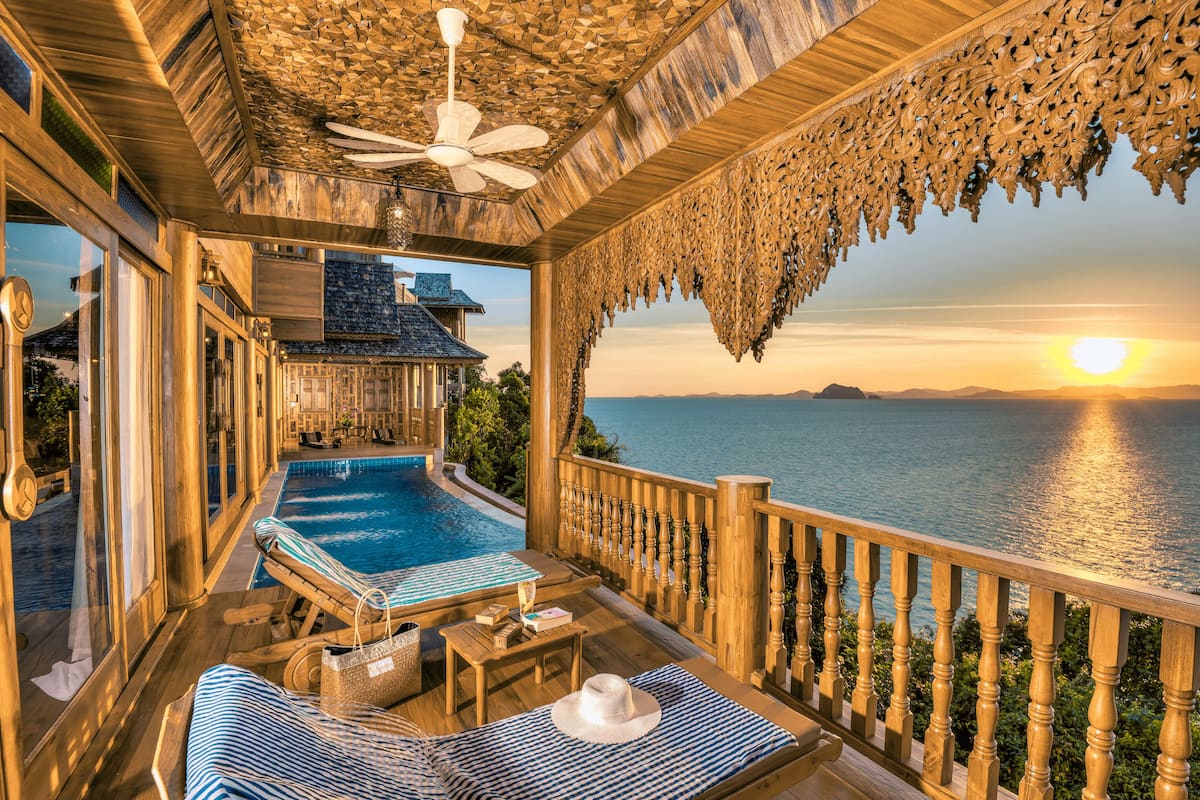 A wooden deck with a pool, lounge chairs, and a view of the sea at sunset.