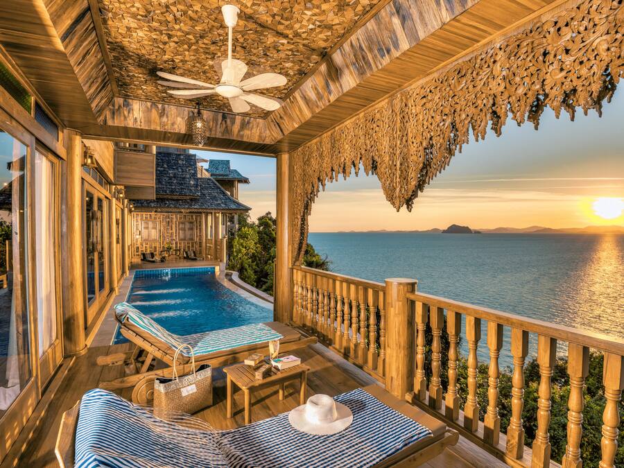 A wooden deck with a pool, lounge chairs, and a view of the sea at sunset.