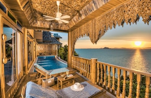 A wooden deck with a pool, lounge chairs, and a view of the sea at sunset.